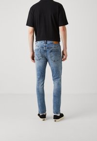 Vaqueros de mezclilla azul claro con ajuste cónico, dos bolsillos traseros y una etiqueta de cuero marrón en la cinturilla; combinados con una camiseta negra de manga corta.