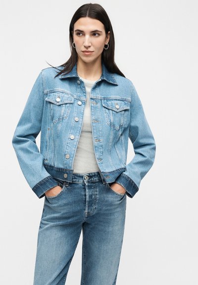 Γυναίκα με μακριά σκοτεινά μαλλιά που φοράει ανοιχτό μπλε τζιν μπουφάν και τζιν παντελόνι, με τα χέρια στις τσέπες, κοιτάζοντας ελαφρώς πλάγια σε απλό λευκό φόντο.