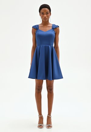 Donna che indossa un abito blu senza maniche con corpetto aderente e gonna svasata, in piedi con sandali argentati con tacco alto e orecchini lunghi.