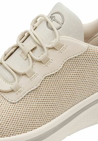 Adidași bej cu un superior din plasă, șireti elastici și un detaliu cu un design ondulat. Talpa este albă, cu o bază texturată pentru aderență.