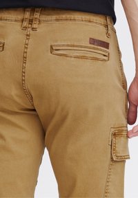 Beige cargobyxor med förstärkta fickor, en läderloggopatch och en slät, hållbar tygtextur.