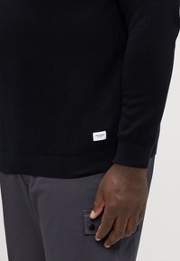 Pulover negru tricotat cu un model texturat, cu manșete și tiv zimțate, având o etichetă mică a mărcii în partea stânga jos. Pantaloni gri închis cu detalii la buzunare.