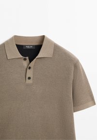 Polo en maille à manches courtes marron clair avec trois boutons noirs et un col, posé à plat sur un fond blanc.