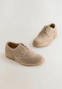 Chaussures beige en daim à lacets avec des détails perforés, bout arrondi et semelle plate en caoutchouc. Fini texturé et accents de couture décoratifs.