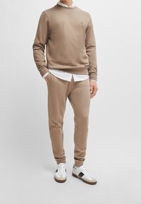 Felpa beige con colletto a girocollo e logo ricamato, abbinata a pantaloni sportivi tapered coordinati, completata da sneakers bianche e beige.