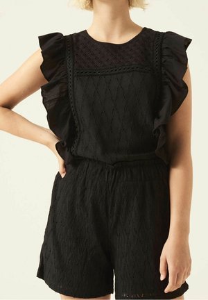 Frau trägt einen ärmellosen schwarzen Textur-Einteiler mit gerafften Schultern und Spitzen-Details, steht mit einer Hand in der Hüfte vor einem schlichten Hintergrund.