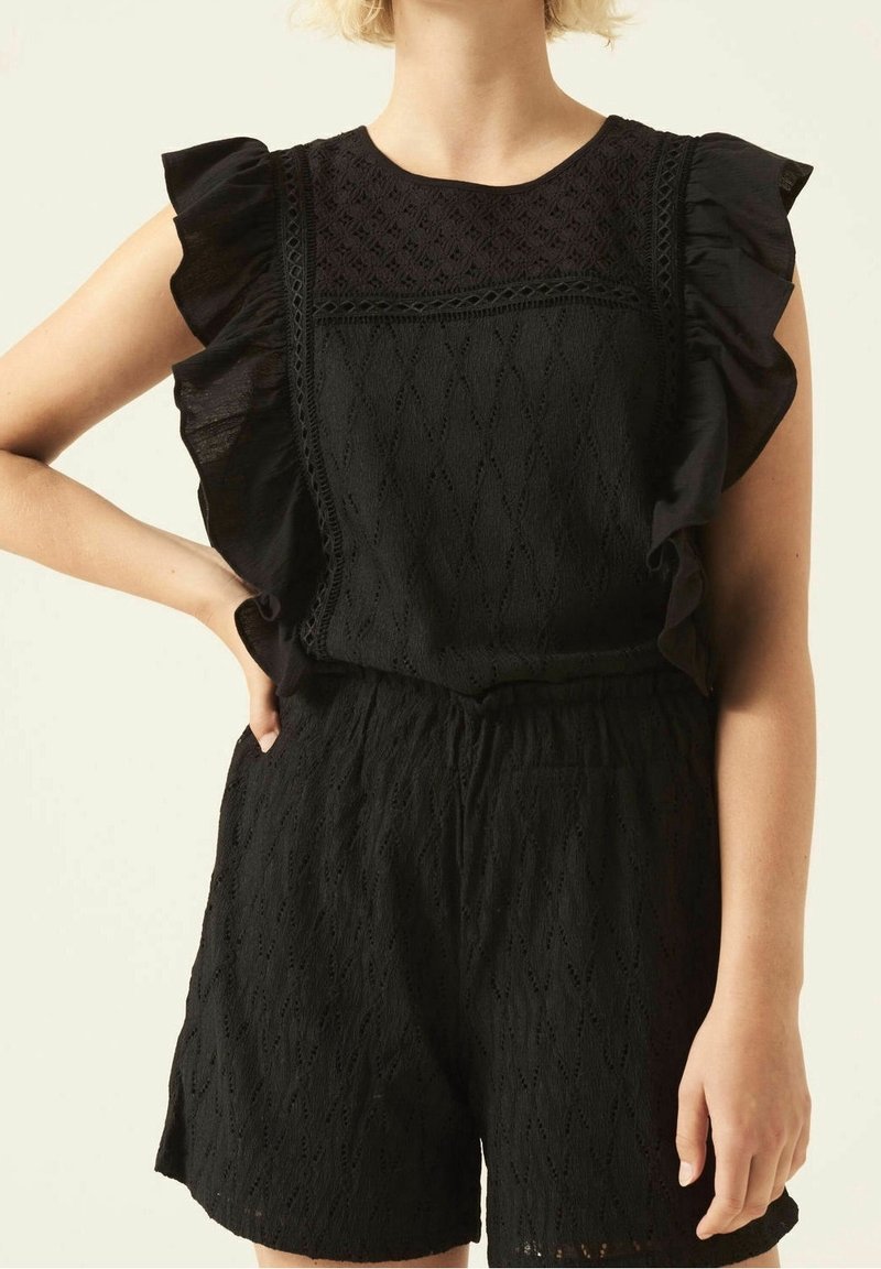 Femme portant une combinaison sans manches noire texturée avec des épaules à volants et des détails en dentelle, debout avec une main sur la hanche sur un fond uni.
