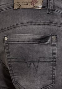 Jeans de mezclilla gris con un bolsillo trasero que presenta costuras en zigzag, acentos de botones de metal y una etiqueta de cuero texturizada.