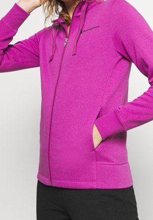 Frau trägt einen knallpinken Champion Kapuzenpullover mit durchgehendem Reißverschluss und Kapuzenbändern, eine Hand in der Tasche, kombiniert mit schwarzen Hosen.
