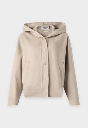 Personne aux longs cheveux foncés porte une veste beige boutonnée avec des manches larges et un pantalon ample vert olive clair sur un fond clair uni.