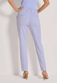 Eine Frau steht mit den Händen in den Hüften, trägt eng anliegende, helllila Hosen und beigefarbene High-Heels vor einer schlichten Wand.