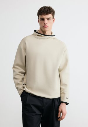 Fiatal férfi, aki bézs színű, fekete szegéllyel díszített laza kapucnis pulóvert és fekete nadrágot visel, egyszerű fehér háttér előtt áll.