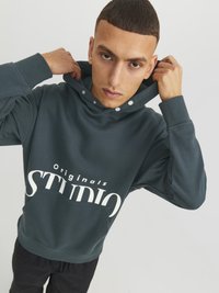 Mørkegrøn hættebluse med stor hvid "STUDIO" trykt. Har en frontlomme og trykknapper på hætten. Glat tekstur.