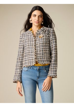 Kvinde iført en frynset sort-hvid ternet jakke med en blomsterbrosje, guld top, øreringe i ringform og blå jeans mod en beige baggrund.