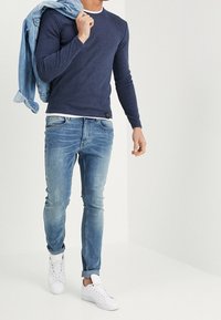 Homme portant un pull marine, un jean bleu clair et des baskets blanches, tenant une veste en jean sur l'épaule en marchant.