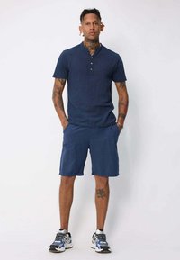 Chemise henley bleu marine à manches courtes avec trois boutons, associée à un short bleu marine assorti. Les chaussures sont des baskets grises et bleues avec un motif.