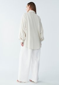 Camicia oversize crema con colletto e maniche lunghe e ampie, abbinata a pantaloni bianchi a gamba larga e sandali con tacco minimalisti.