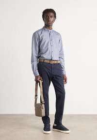 Világoskék gombos ing, sötétkék nadrág, bézs öv és sötétkék sportcipő. Kézben tart egy kis barna crossbody táskát. Az anyagok sima textúrájúak.