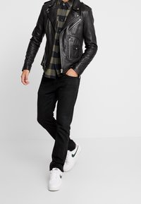 Chaqueta de moto de cuero negro con cremalleras plateadas, llevada sobre una camisa de cuadros verde y negro, combinada con jeans negros y zapatillas blancas.