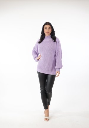 Vrouw staand, gekleed in een lavendelkleurige grofgebreide trui, zwarte leren broek en witte openhielsandalen, tegen een witte achtergrond.