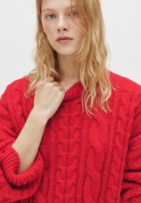 Pulover roșu cu tricotaj în model de cablu, cu guler înalt și croială liberă. Material din lână texturat cu manșete răsucite. Modelul ține ușor gulerul.