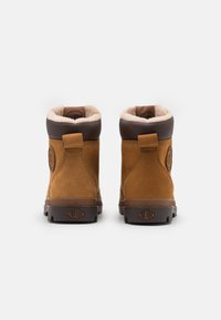 Bottines en suede marron avec une doublure beige douce, bout arrondi et accents marron foncé. Présente un logo sur le côté et une semelle en caoutchouc texturée.