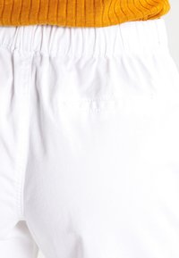 Bílé bavlněné kalhoty s elastickým pasem, které mají jemnou texturu a jednu zadní kapsu. Vrchní díl je ribbovaná hořčicová svetr.