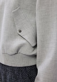 Grijze bomberjack met een gestructureerde stof, ribgebreide zoom en een zijzak met een drukknoopsluiting. Het ontwerp is modern en praktisch.