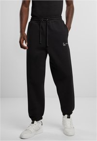 Schwarze Jogginghose mit elastischem Bund, Kordelzug, Seitentaschen und gerippten Bündchen. Mit einem weißen Logo auf dem linken Oberschenkel.