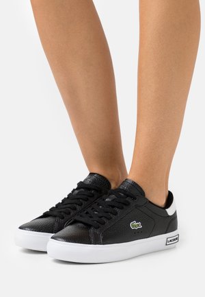 Baskets basses noires Lacoste avec semelles blanches portées pieds nus, montrant un gros plan des jambes et chevilles.