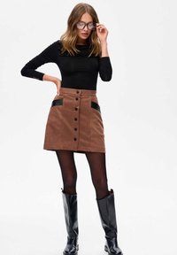 Bruine corduroy rok met zwarte leren look zakken, met een sluiting aan de voorkant met knopen. Gemaakt in combinatie met een zwart langemouwen top en hoge zwarte laarzen.