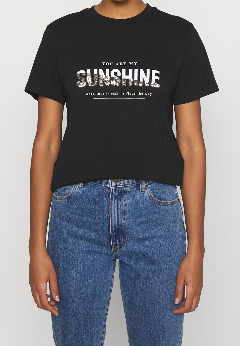 Personne portant un t-shirt noir avec un texte blanc et scintillant disant "Tu es mon soleil, quand l'amour est vrai, il trouve le chemin" et un jean bleu.
