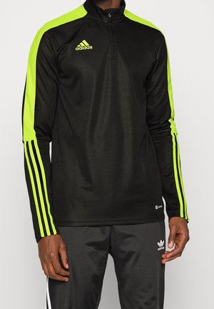 Mężczyzna w czarnej kurtce sportowej Adidas z neonowozielonymi paskami na rękawach oraz dopasowanych czarnych spodniach Adidas z białymi paskami.