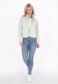Giacca cropped verde chiaro con colletto alto, chiusura con zip e tessuto testurizzato. Indossata sopra una camicia bianca e abbinata a jeans blu e sneaker bianche.
