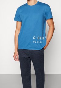 Blå bomulls t-shirt med kort ärm, rund halsringning, och "G-STAR GS E.89" tryckt i ljusgrått på vänster nederkant. Bärs med mörka byxor.