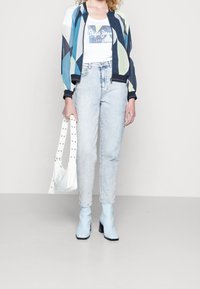 Giacca bomber colorata con motivo geometrico, canotta bianca, jeans a vita alta azzurri e stivali con tacco abbinati. Borsa bianca con borchie.