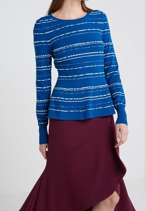 Kobieta w niebieskim topie z długim rękawem z białymi poziomymi przerywanymi paskami w połączeniu z bordową asymetryczną spódnicą.