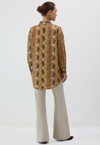 Chemisier à manches longues avec un motif vif jaune, orange et marron. Doté d'un col et de poignets à boutons. Associé à un pantalon large crème.