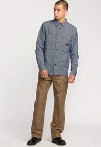 Jasnoniebieska koszula zapinana na guziki z dwoma przednimi kieszeniami i naszywką z logo, w połączeniu z khaki spodniami typu cargo i czarnymi butami. Gładka faktura materiału.