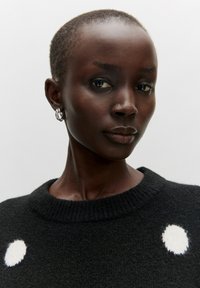Zwarte sweater met een gestructureerde brei, met een wit cirkelpatroon. Model heeft kort haar en zilveren creolen. Neutrale achtergrond.