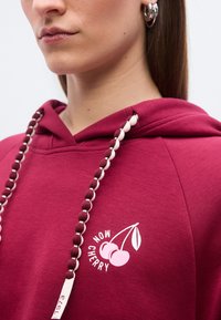 Κοντινό πλάνο ατόμου που φοράει μπορντό φούτερ με κουκούλα, με γραφικό κερασιού και το κείμενο «NOW CHERRY», με κορδόνια τύπου χάντρες και ασημένια κρίκους σκουλαρίκια.