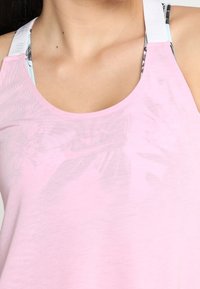 Jasnoróżowy, bezrękawnik o luźnym kroju, z gładką fakturą i białymi, regulowanymi ramiączkami. Zauważalna tropikalna podszewka.