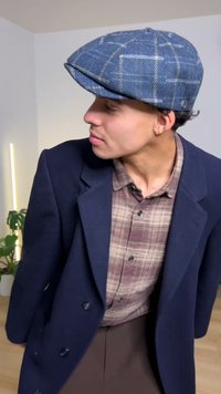 Plaid shirt under a navy coat; gray cap with a checked pattern. Indoor setting with plant and soft lighting.