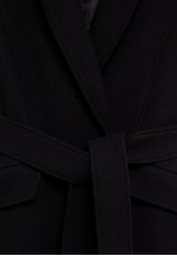 Manteau noir en laine avec de larges revers et une ceinture à nouer à la taille, montrant des coutures détaillées et la texture du tissu.