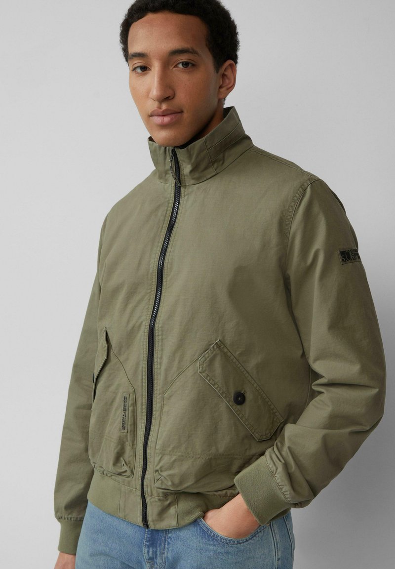 Oliwkowa kurtka bomberka z wysokim kołnierzem, zipem z przodu, dwoma bocznymi kieszeniami oraz ściągaczami na mankietach, wykonana z lekkiego, teksturowanego materiału.