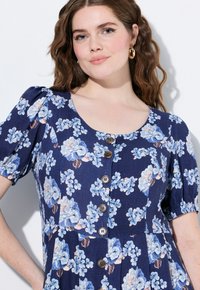 Květinové šaty z navy látky mají modré a růžové květy, krátké nabírané rukávy, výstřih ve tvaru lodičky a hnědé knoflíky vpředu.