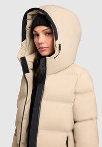 Smėlio spalvos pūkinė striukė su dideliu gobtuvu, juodais akcentais ir užtrauktuku priekyje. Turi kviltinto audinio tekstūrą ir reguliuojamą raištį.