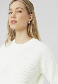 Pull en tricot blanc avec une texture côtelée et un col ras du cou. Présente des coutures minimales et une coupe décontractée. Des boucles d'oreilles en argent complètent le look.