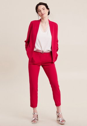 Blazer rouge et pantalon assorti à la coupe ajustée, accompagné d'un haut blanc à bord en dentelle. Des talons blocs argentés complètent le look.