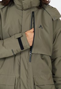 Olivgrön vattentät jacka med dragkedja framtill, justerbara kardborreband vid ärmarna och två stora framfickor. Har en logotyp nära dragkedjan.